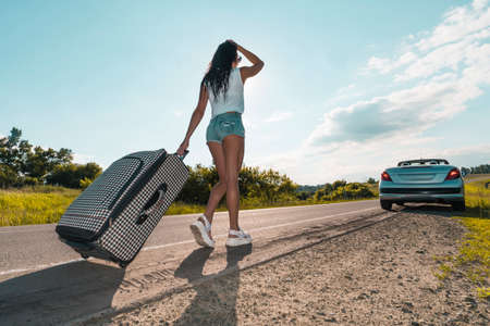 Girl dragging a large heavy suitcase. beautiful young girl in sexy denim shorts with a suitcase is waiting for a passing car to continue traveling. road trip concept. rear view backの写真素材