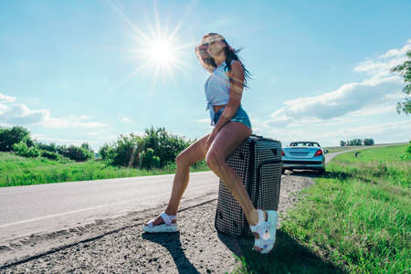 Girl in the bright sun on road with Luggage on the background of the car. caucasian woman sitting on a suitcase. The concept of adventure and travel alone. Road trips. hitchhiking. Summer weekend.の写真素材