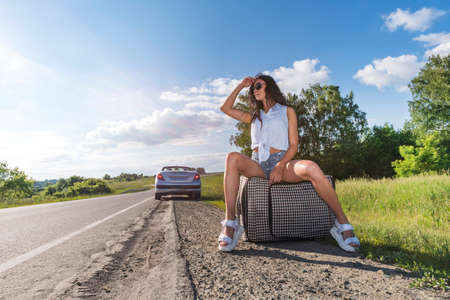 young woman hitch-hiking on a road with luggage bag at the fields. girl is sitting on baggage and waiting for the car to leave the village. concept of road travel and hitchhiking. Towards adventure.の写真素材