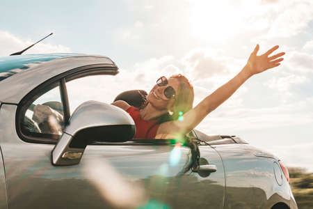 happy young woman enjoying a ride in a convertible car. hand greeting. concept of road travel and adventure. riding with open roof flight effect.の写真素材