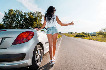 Road scene: sexy brunette girl standing near their broken car and hitchhiking. Rear view. ran out of gas. Problems with cars on the road. Broken car. girl raises her finger. country roadの写真素材