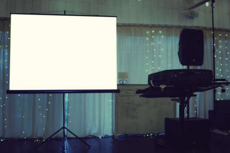 Empty projection screen put in the centre of a illuminated stage in a banquet hall. Projection screen equipment at the decorated curtain background. Facilities for video projection at a festive eventの写真素材