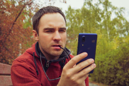 man thinking curiously, holding a smartphone, sitting outdoor in garden. A young guy looks indignantly at the screen of his smartphone. Reading bad news. solving business problems onlineの写真素材
