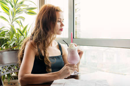 Young beautiful girl drinking a strawberry-flavored milkshake in a restaurant on a window background in daylight. The girl holds a glass of non-alcoholic cocktail.の写真素材