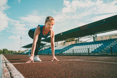 Ready to go. Young fitness blonde girl wearing blue top, black tights, sneakers and pony-tail on the starting line of stadium track, preparing for a run. Healthy lifestyle concept.の写真素材