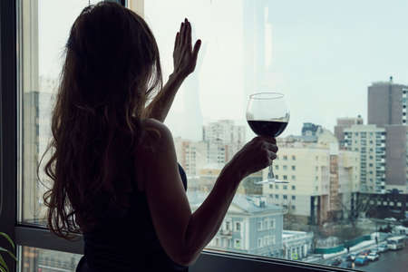 single luxury beautiful woman in black dress with wine standing near window looking aside. Lonely young woman at home drinking alcohol. Female alcoholism.の写真素材