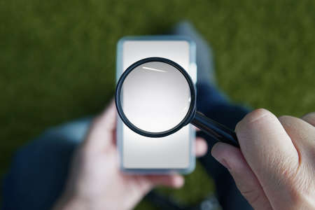 Close up of male hands holding smartphone with magnifying glass above the device screen. Shot of a sitting person with mobile in one hand and the loupe in the other. Concept og mobile searchingの写真素材