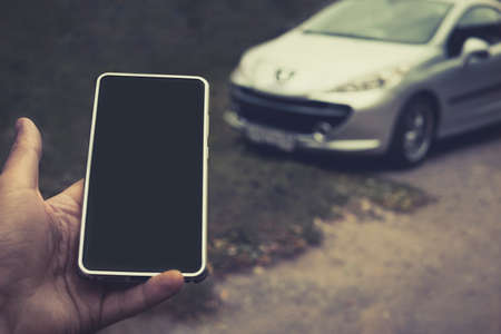 Smartphone with black screen in male palm at parked vehicle background. Engine start mobile service. Phone sends signals to unlock the vehicle. Usefull car application. Integration of car and phoneの写真素材