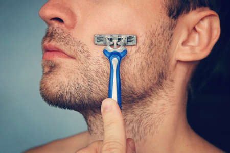 view of a young bearded man with a brutal razor. face on the blue background. big rough hairy bristles on the skin. side view close upの写真素材
