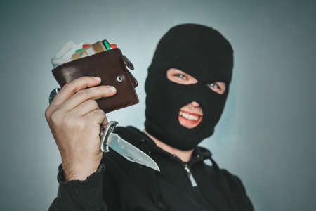Masked criminal at the black background grins looking at the viewer and showing a purse in hand....