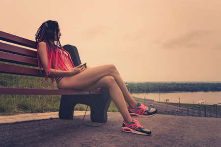 Fashionable woman posing at street and sitting on bench with cell phone and headphones. Fashion girl listening music heavy metal rock on mobile.の写真素材