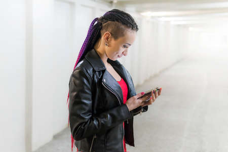 young punk style lady in black jacket with an unusual hairstyle looks into screen of your smartphone against tunnel background with bright light at the end.の写真素材