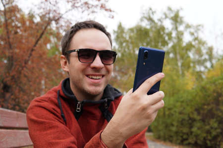 Young man sitting at park and using smart phone. Happy smiling guy reading a message on cellphone while sitting on a bench in a park. man surfing net with smartphoneの写真素材