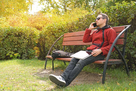 Happy casual man with laptop and take away coffee talking on mobile. Handsome male freelancer working outdoors in the park. Technology, communication, education and remote working concept, copy spaceの写真素材