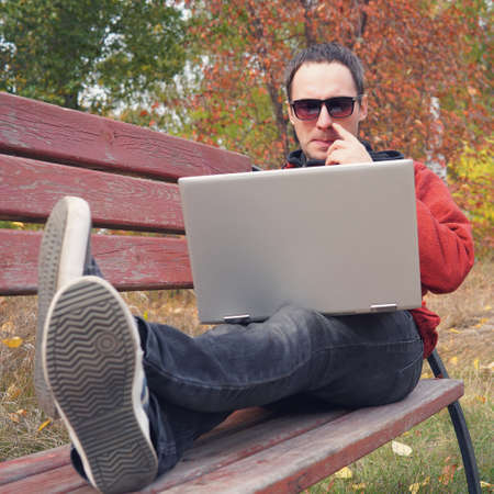 Young male student programmer work study outdoors. Programmer developer works with his laptop in the Park outside the office. Pensive man with an ultrabook on a benchの写真素材