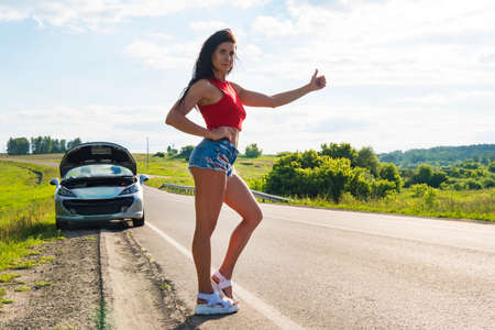 Beautiful woman hitchhiking by a broken car. girl stands at his car and waiting for help. beautiful young sexy girl in short shorts stands at her convertible. Problem car on road. ran out of gas.の写真素材