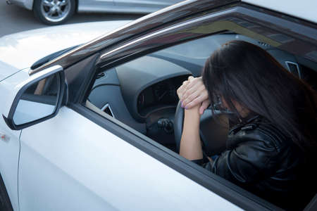 View form front window to a stressed or tired brunette girl in car lying on steering wheel. Female driver resting in vehicle. Young girl felt sleepy or sick while driving. Woman leans car wheel.の写真素材