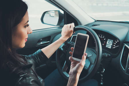 Young woman looking to her smartphone while driving car at front window background. Distracted female driver. Concept of dangerous driving. Woman driving and holding cellphone with blank screenの写真素材