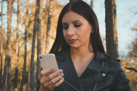 Close up of attractive brunette girl holding smart phone at unfocused forest background. Female cant stop checking her cellphone even walking in park. Young woman using phone in woods. Connection.の写真素材