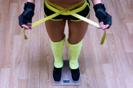 Top-down view to fitness woman standing on scale and taking waist measurements with metric tape. Concept of losing weight in gym. Girl standing on scales with written word Help on display. Shaping.の写真素材
