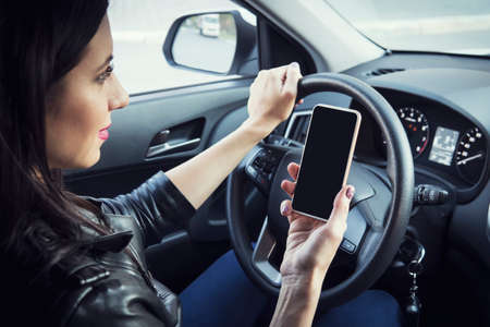 Side view of young woman holding blank screen cell phone while driving car. Female driver hand on steering wheel and checking out her smartphone in moving vehicle. Dont text and drive concept.の写真素材