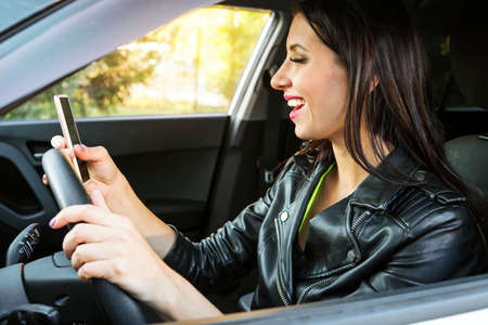 Happy smiling woman holding her smart phone sitting inside a car. Female driver laughing reading message and chatting in mobile. Girl looks at her cellphone screen and smiles while being at wheel.の写真素材