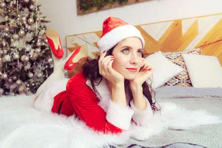 young beautiful girl in a red Santa Claus suit is lying on bed and dreamily looking into the distance against background of apartment decorated for Christmas.の写真素材