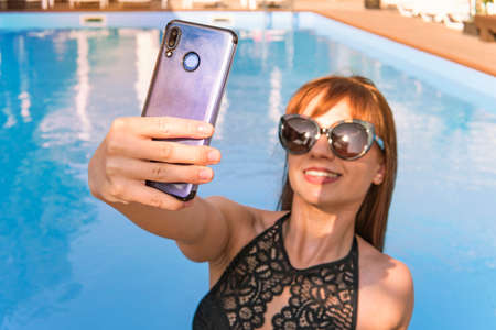 Vacation and technology. Colorful portrait of pretty young woman taking selfie portrait near swimming pool. Selfie from the smartphone of a young young girl in a black bathing suitの写真素材