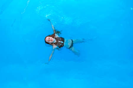 Attractive woman in a black bathing suit floating on her back in the swimming pool and relaxing,...