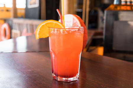 Non-alcoholic cocktail orange in a glass with a slice of orange on the edge with pieces of ice on the bar in the cafe. A glass of red juice on a wooden tableの写真素材