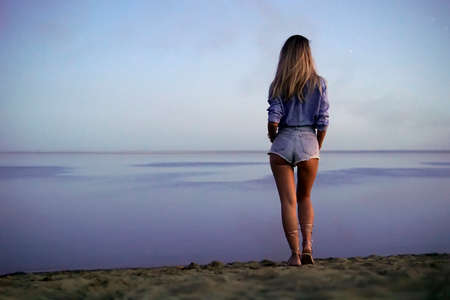 a young beautiful sexy girl stands on the lake and looks into the distance at the horizon in evening. rear view. Beautiful serene nature. minimalism. blonde walks on the sand. copy spaceの写真素材