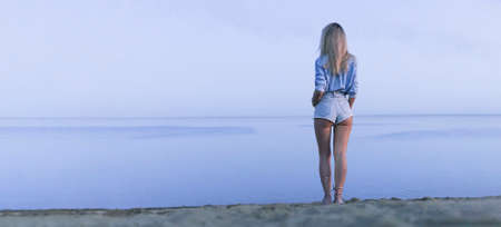 a young beautiful sexy girl stands on lake and looks into the distance at the horizon in evening. view from the back. Beautiful serene nature. minimalism. blonde walks on the sand on beach in morningの写真素材