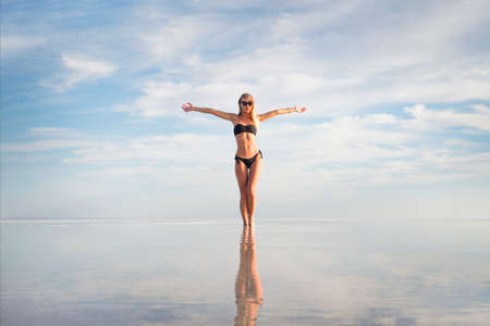 Young adult woman in black white swimsuit standing in water at water texture. Cute sexual girl on summer lake background. hands to side. Low white clouds reflected in clear water. Bolivia Uyuniの写真素材