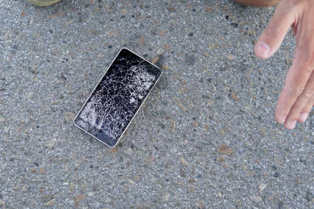 man is sitting and holding a broken smart phone with a cracked screen. guy dropped his mobile phone on the asphalt and broke it. A frustrated man with a broken cell phone.の写真素材