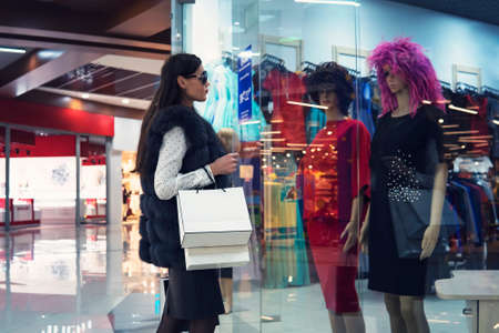 Young woman in shopping centre standing near the shop window and looking at two mannequins in dresses and wigs. Trendy attractive girl is doing shopping in mall. Lady in glasses looking for outfit.の写真素材