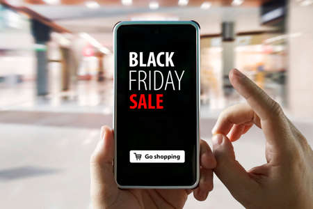 Hands holding mobile phone with Black Friday sale on screen on blurred image of shopping mall. Purchase goods via the Internet in the mobile application. Concept promotion sale discount .の写真素材