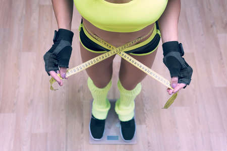 Top-down view to fitness woman standing on scale and taking waist measurements with metric tape. Concept of losing weight in gym. Girl standing on scales with written word Help on display. Shapingの写真素材