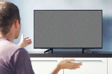 Frustrated Man Sitting On Sofa In Front Of Television With No Signal. young guy emotionally watching TV and waving his hands in discontent.の写真素材