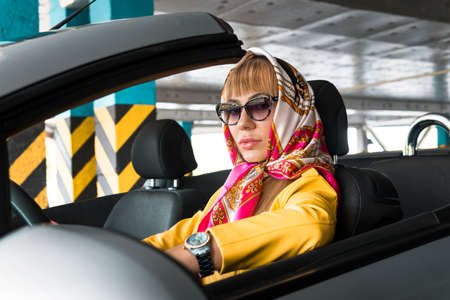 Young woman in sunglasses, in a headscarf driving convertible car. Blond fashion model indoor...