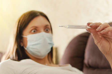 Girl in a mask from a virus with thermometer in hands. Temperature, thermometer. woman is surprised by high temperature on thermometer. Fear of getting sick. emotion bewilderment. Coronovirus epidemicの写真素材