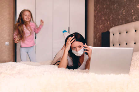 Children make it difficult to focus on work. mother in protective mask working from home during quarantine time at coronavirus. Child at home, kindergarden is closed. sick woman works under quarantineの写真素材