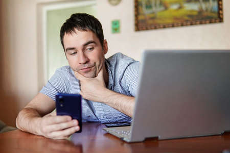 Businessman working on laptop in home office. Connecting your smartphone to your computer via Wifii. The guy works remotely against the background of the home environment.の写真素材