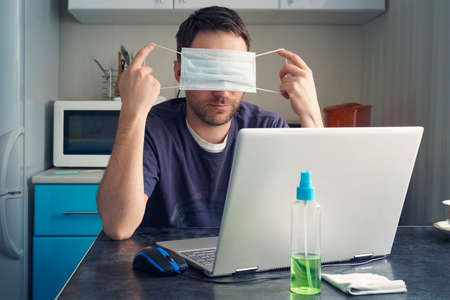 A man is sitting at a table and working on a laptop in home clothes against the background of the kitchen. Tired of remote work and quarantine, the guy removes the protective mask from his face.の写真素材