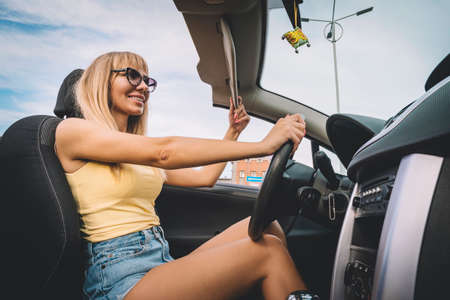 Young cute girl preens while driving. blonde admires herself in the mirror. Happy female driver behind the wheel. woman lowers the sun visor holding the steering wheelの写真素材