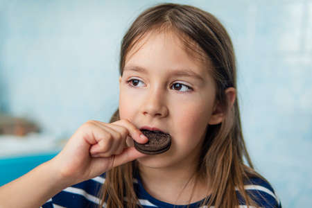 Close up of a girl eating a round chocolate cookie sitting in the kitchen at home. Harmful but delicious food. Sweetness.の写真素材