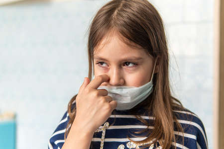 Toddler girl with medical mask on face. Covid19 corona virus outbreak. girl touches face dirty hands, it's not safe. Sad girl is tired of sitting at home on self-isolation quarantine during pandemic.の写真素材