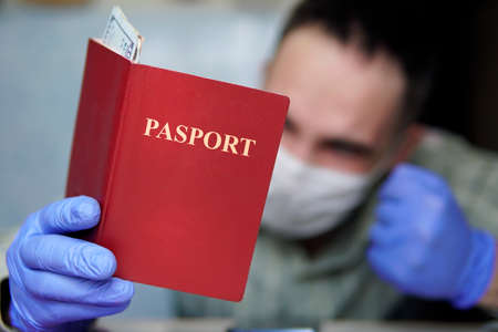 A bored tourist is quarantined at home, wearing a mask and gloves, looking at his passport and...