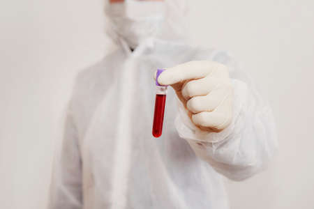 a lab technician in a protective suit and mask holds an ampoule full of blood for testing for viruses or disease. DNA test. Hand holding a blood sample for analysis. white background,の写真素材