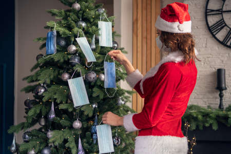A young woman in a red Santa Claus costume decorates the Christmas tree with medical masks. The...