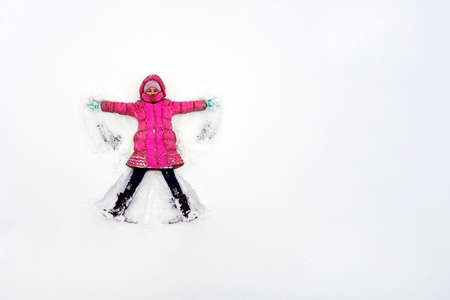 A young child playing in the snow making an angel. A girl in a red jacket plays in the snow in...